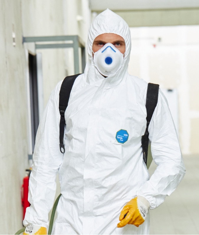 Teaserbild FORTIS Atemschutz - Eine Person in einem weißen Schutzanzug mit Kapuze, FORTIS Atemschutzmaske, gelben Schutzhandschuhen und schwarzem Rucksack läuft durch einen hellen Flur