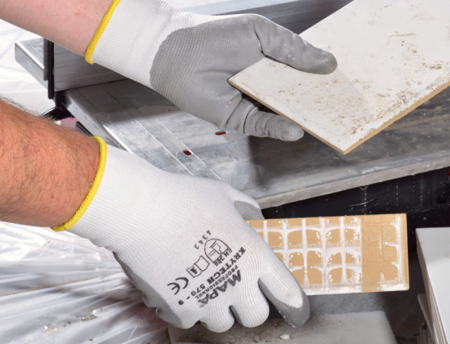 Zwei Hände in weißen MAPA Sicherheitshandschuhen halten und bearbeiten Fliesen an einer Schneidemaschine