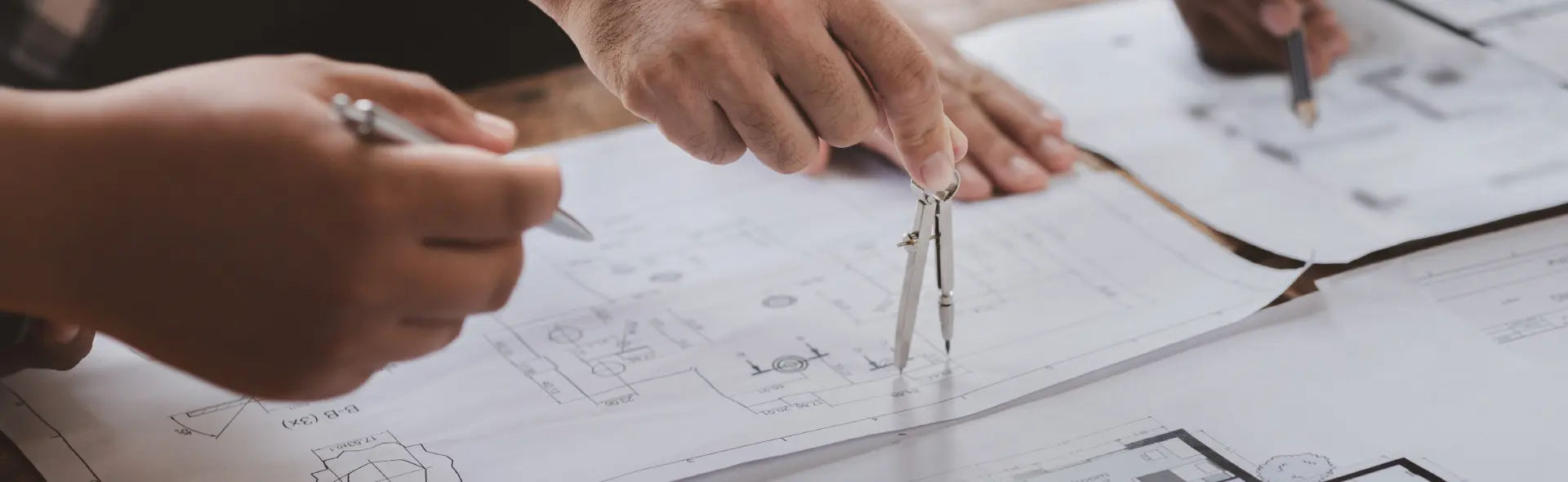 Mehrere Hände arbeiten gemeinsam an technischen Zeichnungen. Eine Hand hält einen Zirkel, während andere Hände Stifte verwenden, um auf den Plänen zu zeichnen oder zu notieren. Die Szene vermittelt Teamarbeit und Präzision in der Planung.