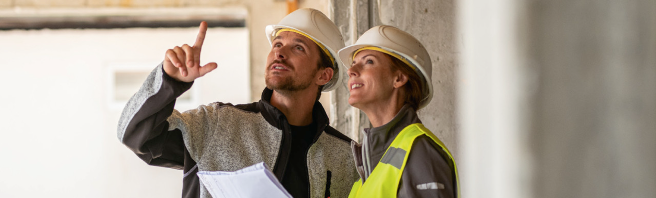 Zwei Personen in persönlicher Schutzausrüstung von FORTIS stehen auf einer Baustelle. Eine Person zeigt mit dem Finger nach oben, während die andere Person ein Dokument in der Hand hält und aufmerksam zusieht.