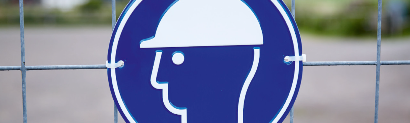 Rundes, blaues Hinweisschild mit weißem Symbol eines Kopfes mit Schutzhelm, angebracht an einem Bauzaun. Das Schild weist auf die Pflicht zum Tragen eines Schutzhelms hin.