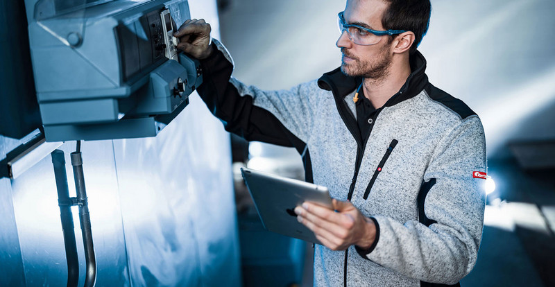 Eine Person in persönlicher Schutzausrütstung von FORTIS bedient ein technisches Gerät an der Wand und hält dabei ein Tablet in der anderen Hand