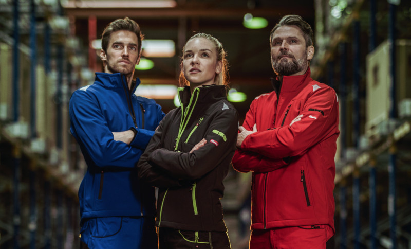 Drei Personen in farbiger persönlicher Schutzausrütstung von FORTIS stehen mit verschränkten Armen nebeneinander in einem Lager oder einer Industriehalle und schauen in die Ferne.