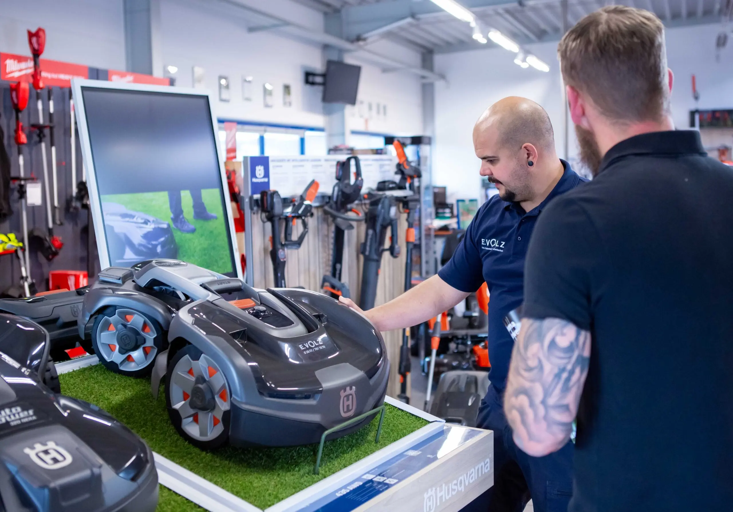Zwei Personen betrachten einen schwarzen Husqvarna-Rasenmähroboter, der auf einer Ausstellungsfläche präsentiert wird. Im Hintergrund sind weitere Gartengeräte und ein Bildschirm mit einer Produktpräsentation zu sehen.