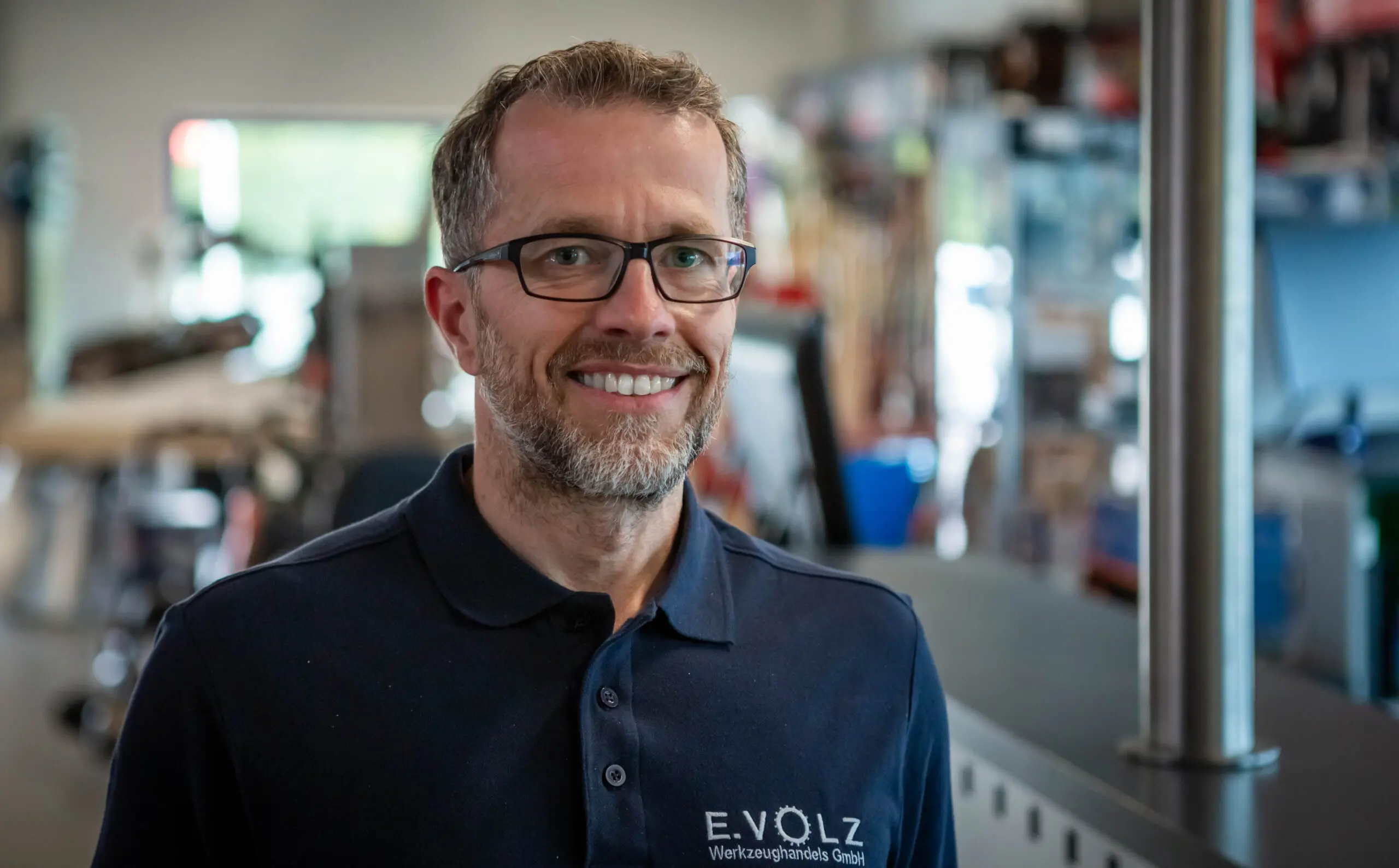 Eine Person trägt ein dunkelblaues Poloshirt mit dem Logo „E.VOLZ Werkzeughandels GmbH“ in einer Werkstattumgebung. Im Hintergrund sind Regale, Werkzeuge und eine helle Fensterfront zu sehen.