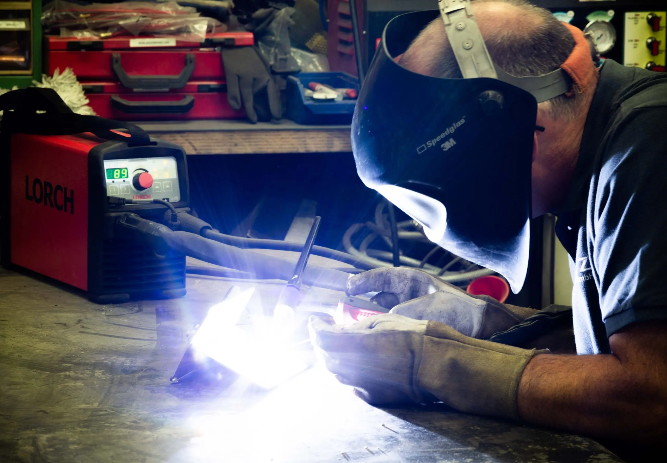 Eine Person schweißt Metall mit einer roten Schweißmaschine der Marke „Lorch“. Sie trägt eine Schweißmaske und Schutzhandschuhe. Der Arbeitsplatz ist mit Werkzeugen und einem Werkbankbereich ausgestattet, während helles Licht vom Schweißvorgang ausgeht.