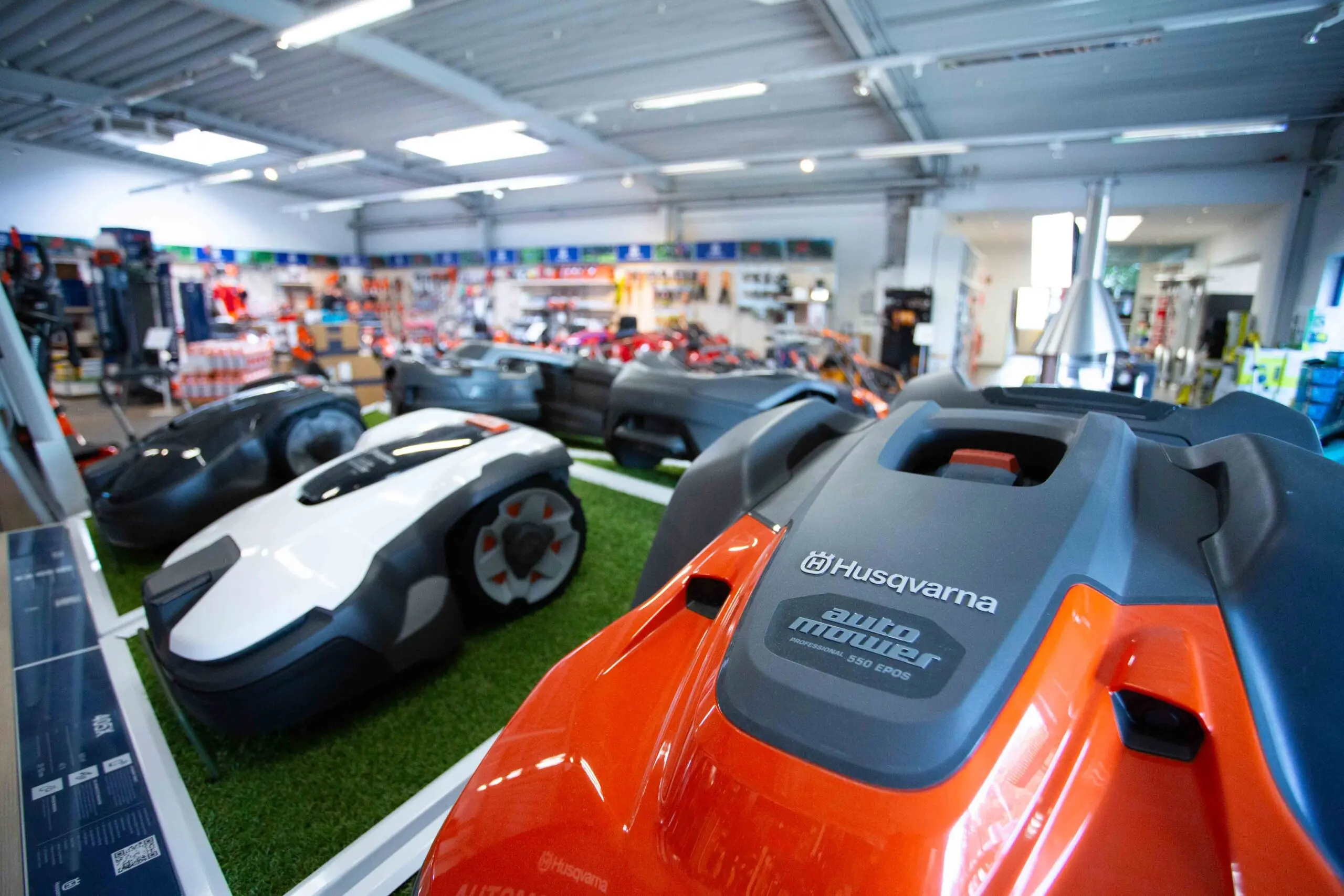 Mehrere Husqvarna Automower-Rasenmähroboter, darunter ein orangefarbener „Automower 550 EPOS“, werden auf einer Ausstellungsfläche mit künstlichem Rasen präsentiert. Im Hintergrund ist eine gut ausgestattete Werkstatt mit verschiedenen Gartengeräten und Werkzeugen zu sehen.