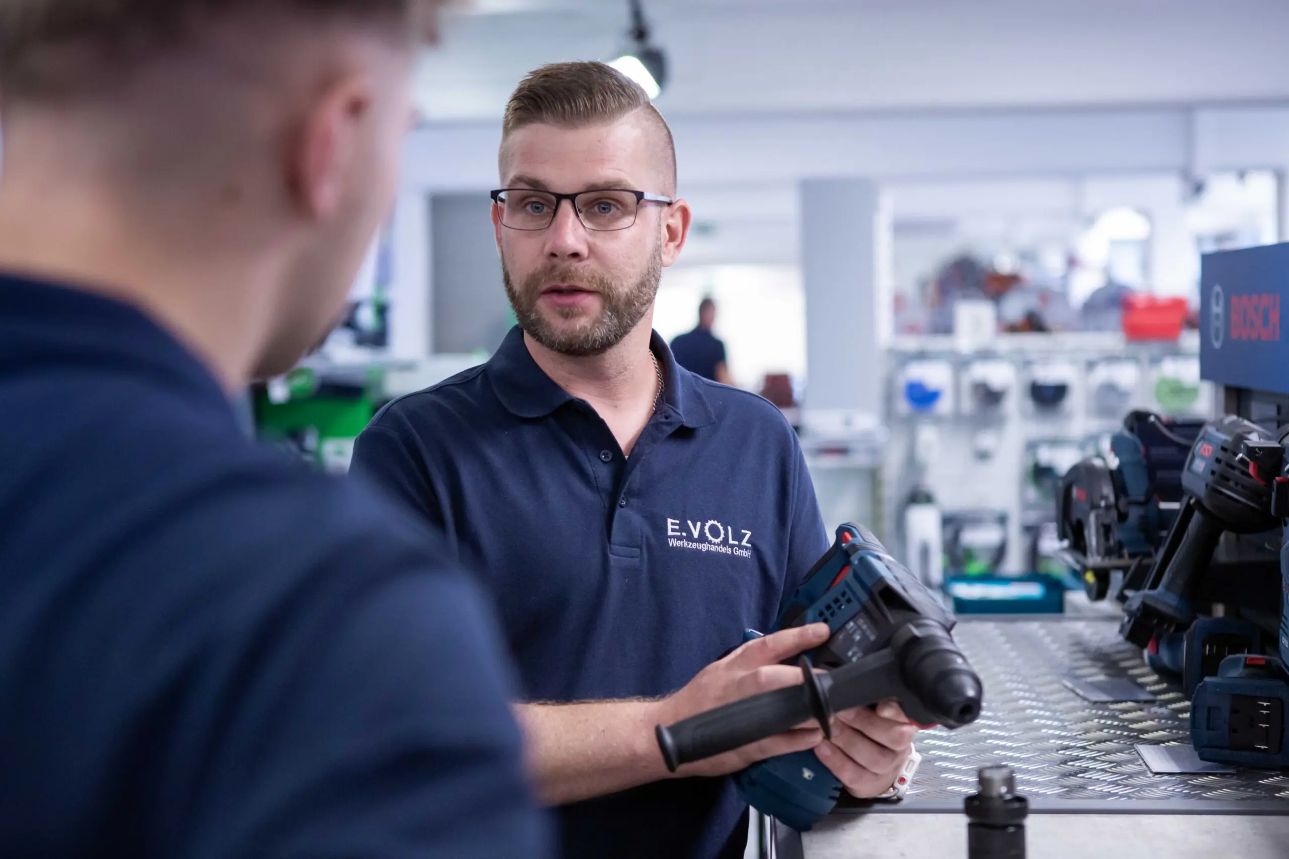 Zwei Personen in einer Werkstatt, eine Person hält eine Bohrmaschine und erklärt deren Funktionen. Beide tragen dunkelblaue Poloshirts mit dem Logo „E.VOLZ Werkzeughandels GmbH“. Im Hintergrund sind Regale mit Werkzeugen und Maschinen zu sehen.