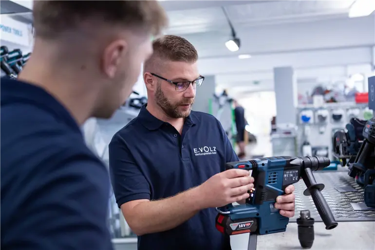 Zwei Personen in einer Werkstatt, eine Person hält eine blaue elektrische Bohrmaschine mit rotem Griff und erklärt deren Funktionen. Beide tragen dunkelblaue Poloshirts mit dem Logo „E.VOLZ Werkzeughandels GmbH“. Im Hintergrund sind Werkzeuge und Arbeitsgeräte zu sehen.