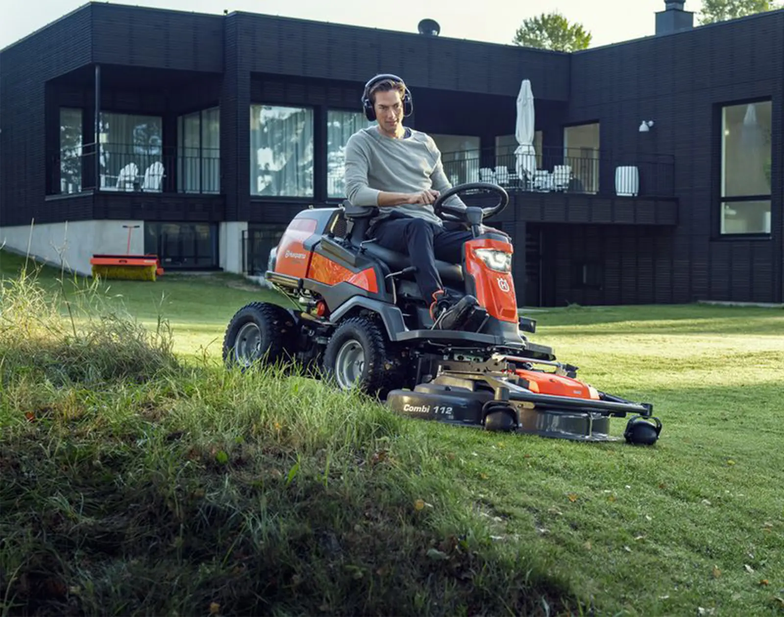 Eine Person fährt einen orangefarbenen Aufsitzrasenmäher auf einer Grünfläche. Im Hintergrund ist ein modernes Gebäude mit großen Fenstern und einer Terrasse zu sehen. Der Rasenmäher ist mit einem „Combi 112“-Mähwerk ausgestattet.