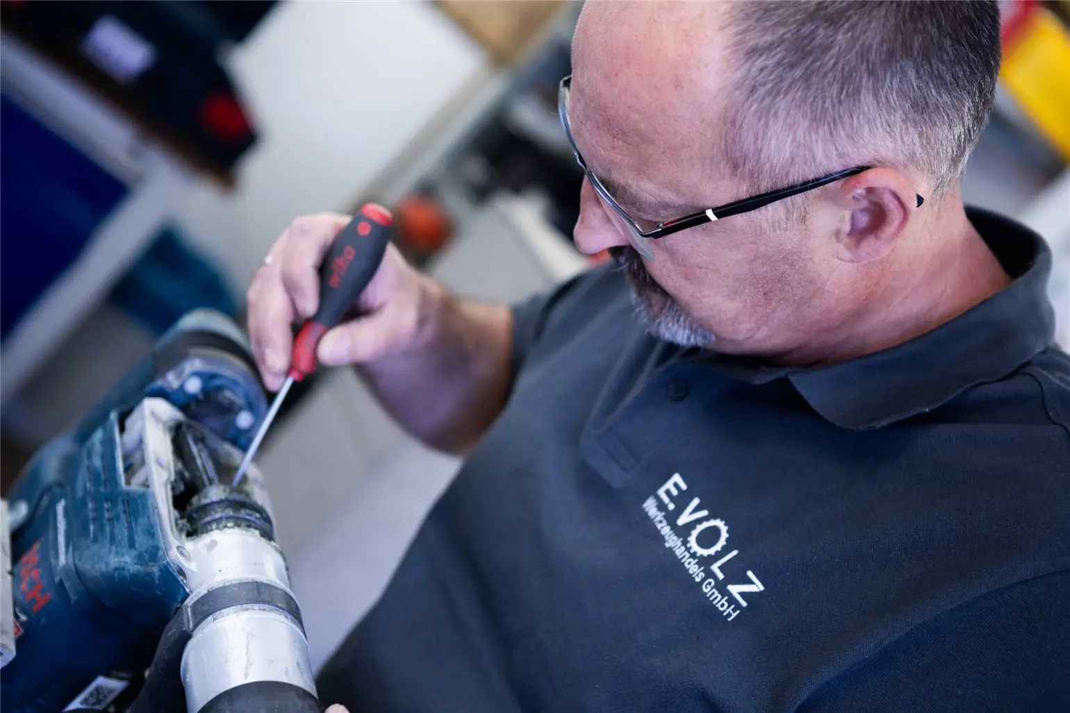 Eine Person arbeitet mit einem Schraubendreher an einer blauen elektrischen Bohrmaschine. Sie trägt ein dunkelblaues Poloshirt mit dem Logo „E.VOLZ Werkzeughandels GmbH“. Im Hintergrund sind unscharf Werkzeuge und Regale zu erkennen.