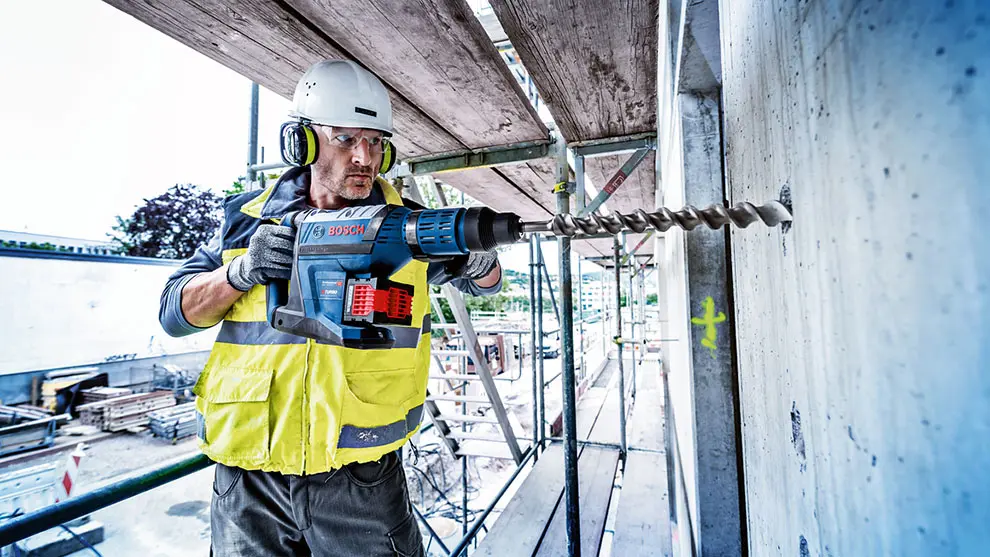 Eine Person arbeitet mit einer blauen „Bosch“-Bohrmaschine und bohrt in eine Betonwand. Sie trägt einen weißen Schutzhelm, Gehörschutz und eine gelbe Warnweste. Im Hintergrund sind Baugerüste und eine Baustelle zu sehen.