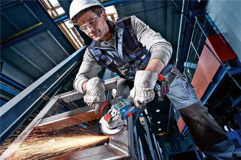 Ein Arbeiter schleift mit einem blauen „Bosch“-Winkelschleifer Metall, wobei Funken sprühen. Er trägt Sicherheitsausrüstung, darunter einen weißen Schutzhelm, Handschuhe und einen Sicherheitsgurt. Die Szene spielt in einer industriellen Umgebung mit Stahlträgern und hohen Decken.