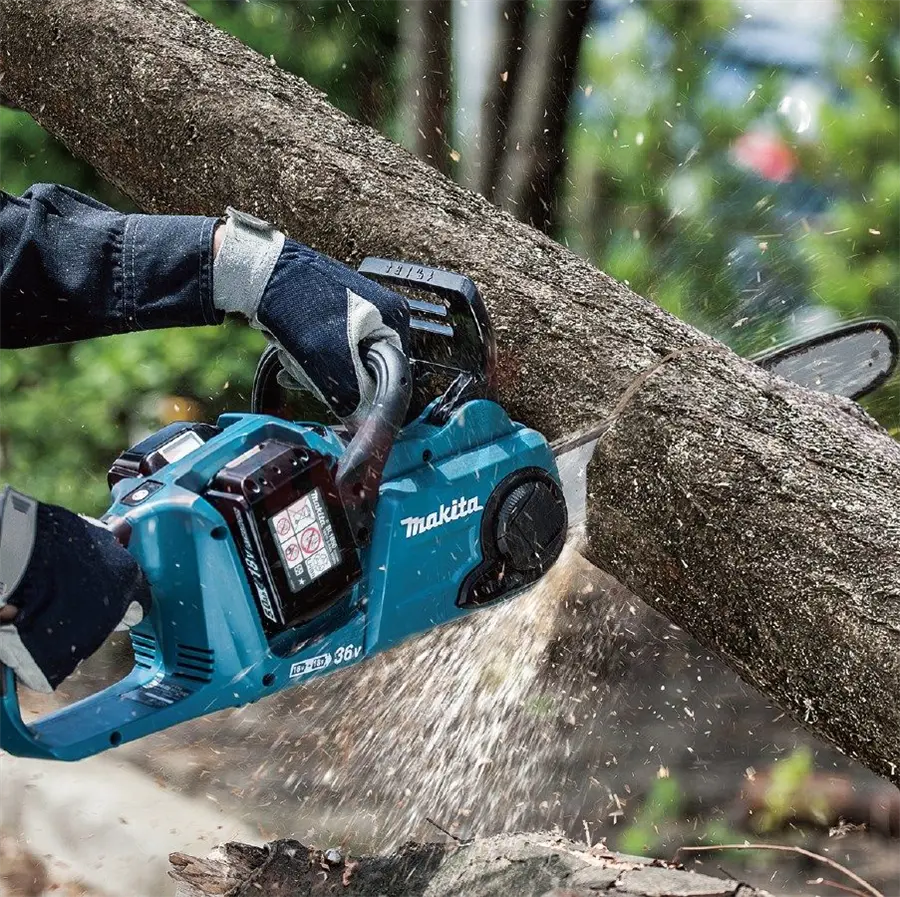 Eine blaue „Makita“-Akku-Kettensäge schneidet durch einen dicken Baumstamm, während Holzspäne sprühen. Die Person trägt Arbeitshandschuhe und arbeitet draußen, umgeben von Bäumen und Grün.