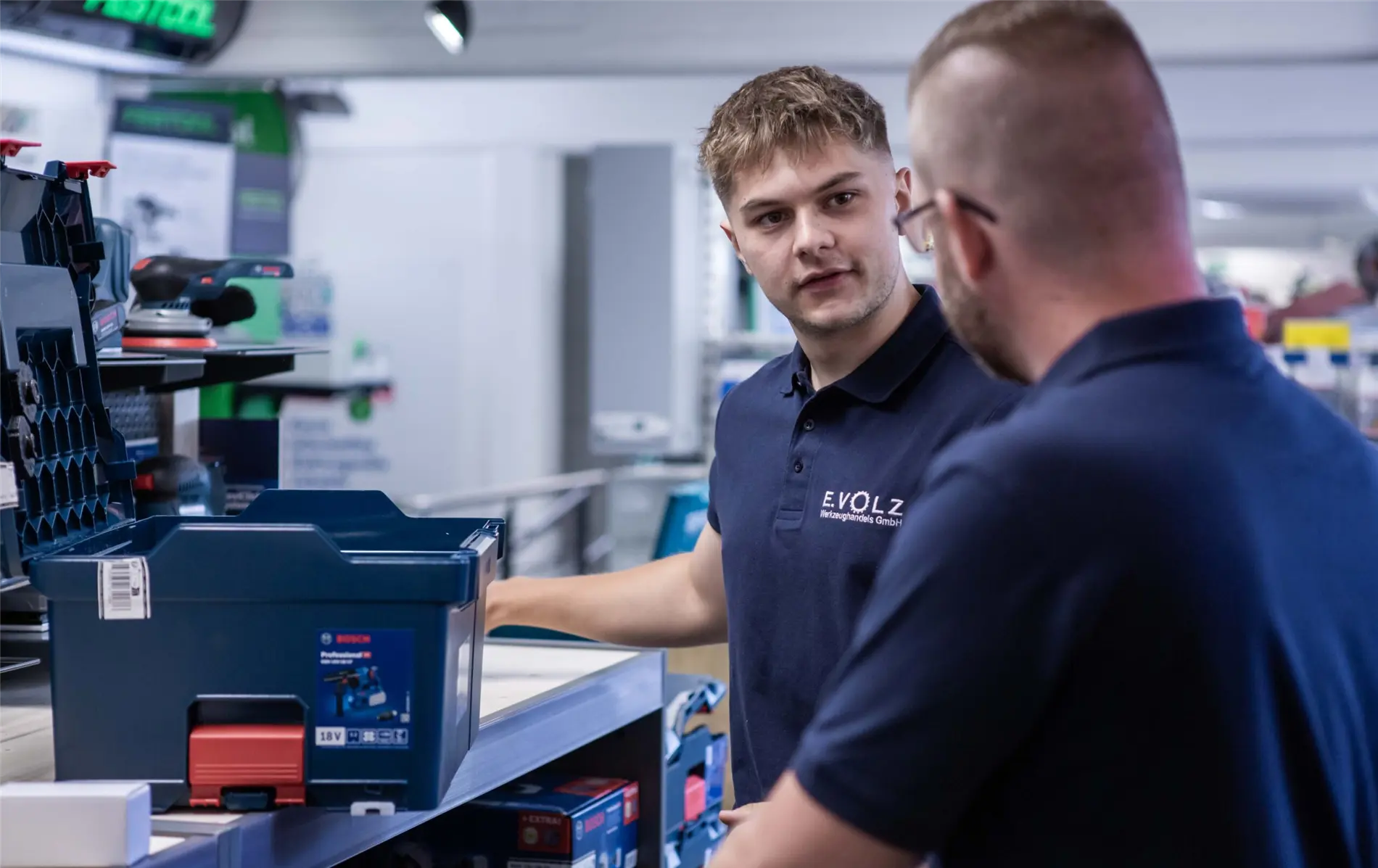 Zwei Personen in einem professionellen Gespräch, beide tragen dunkelblaue Poloshirts mit dem Logo „E.VOLZ Werkzeughandels GmbH“. Im Hintergrund ist eine Werkstatt mit Werkzeugen und Maschinen zu sehen. Der Text „Vertrauensvolle Zusammenarbeit“ sowie weitere Informationen über hochwertige Handwerksarbeit und Experten für Profi-Werkzeug sind eingeblendet.